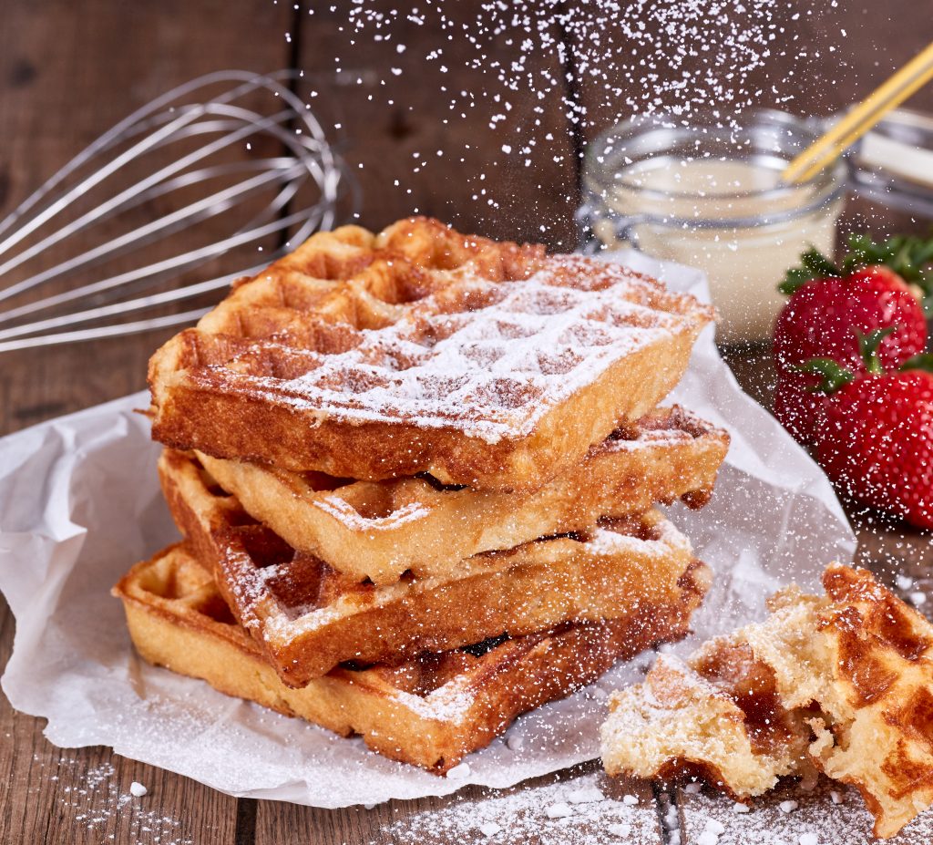 Culinaria süße Waffel, vier Stück gestapelt, von oben fällt Puderzucker auf die Waffeln