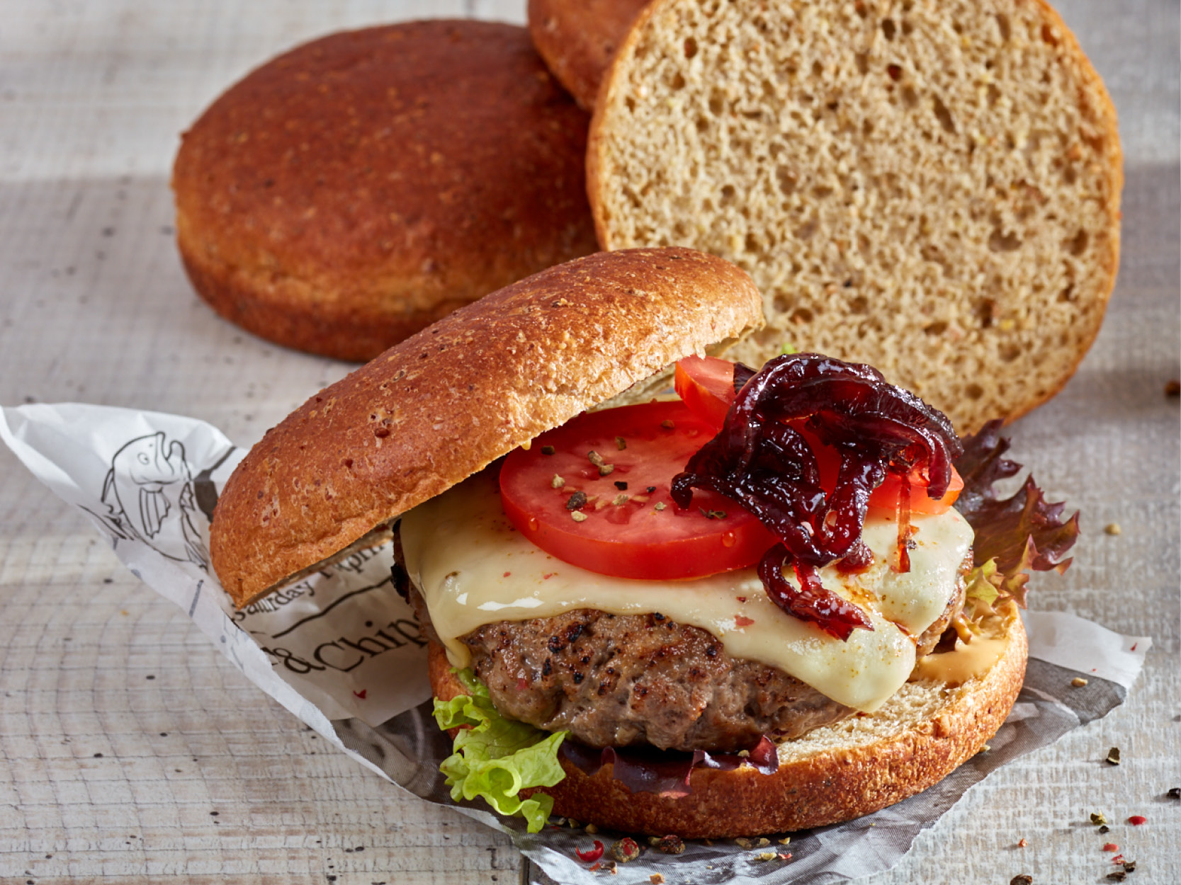 Protein Burgerbun. Im Vordergrund ein belegter Burger mit Rinderpatty, Käse, Tomate und gerösteten Zwiebeln